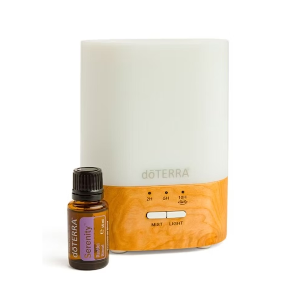 A doTERRA essential oil diffuser with a white upper body and a light wood-grain lower base, shown next to a small amber bottle of doTERRA Serenity essential oil on a white background.
