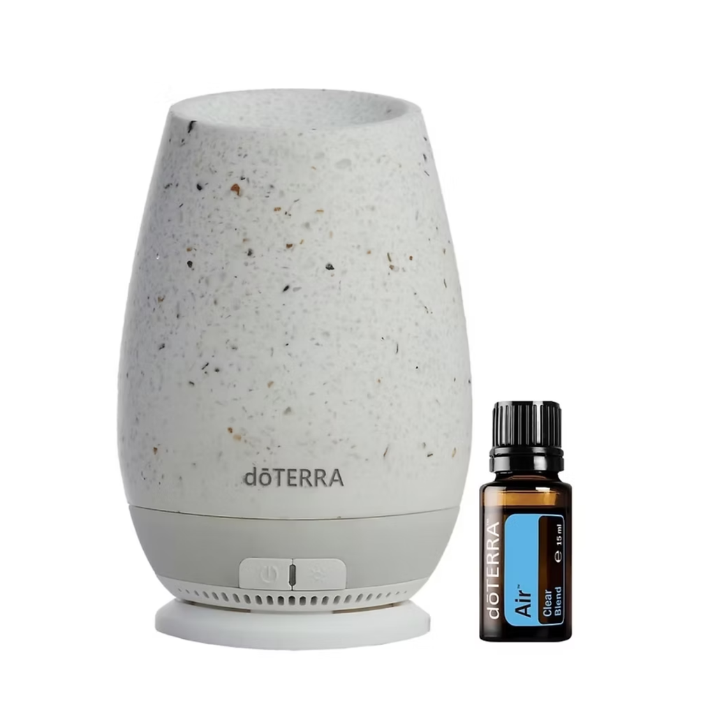 A doTERRA Air diffuser lamp in a speckled off-white ceramic style next to a small amber glass essential oil bottle with a black cap and blue label, on a plain white background.