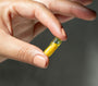 A close-up of a hand holding a small translucent yellow capsule pill between the fingers against a dark gray background.