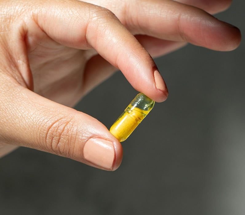 A close-up of a hand holding a small translucent yellow capsule pill between the fingers against a dark gray background.