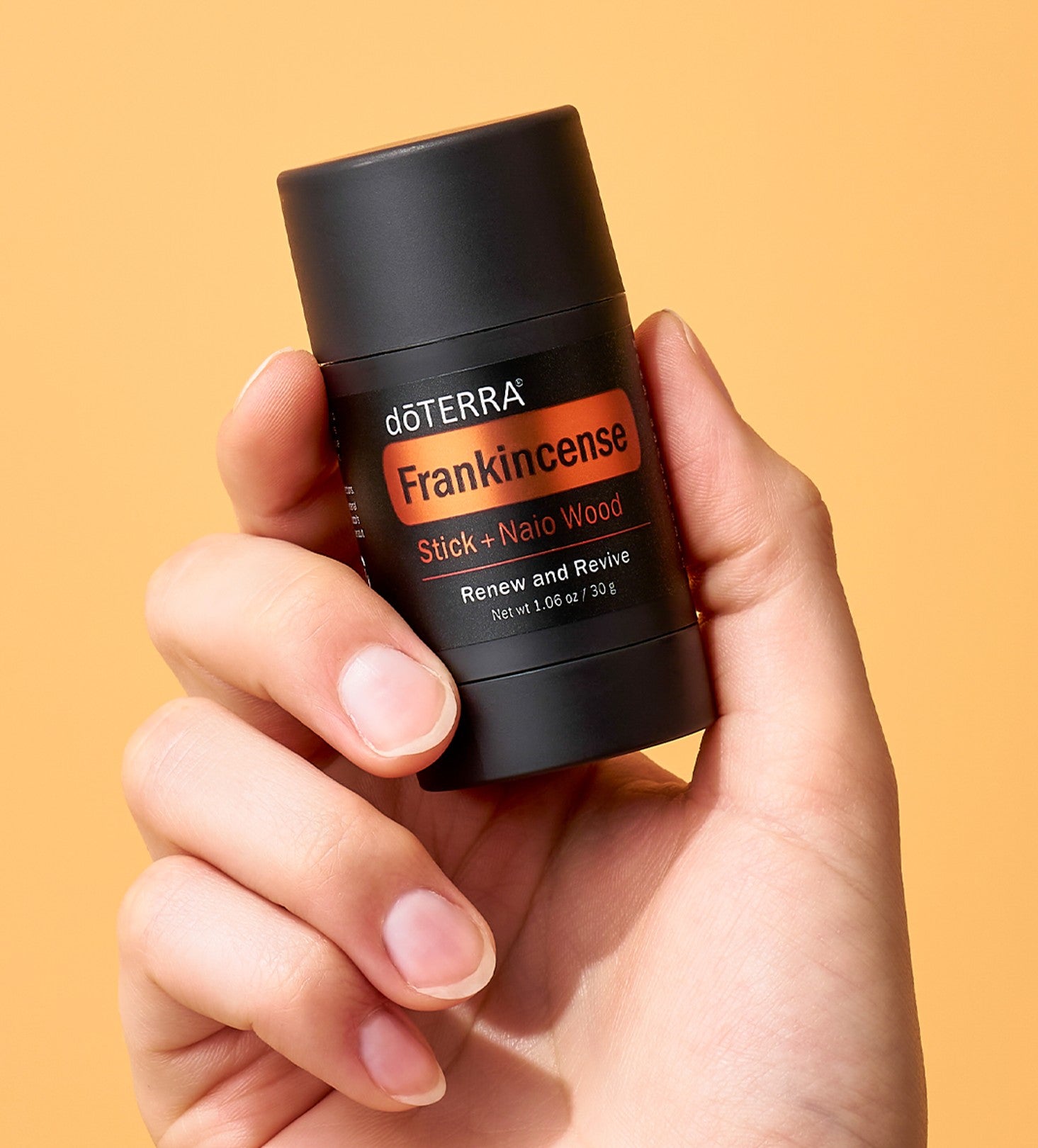 A hand holding a black doTERRA Frankincense Stick + Alamo Wood deodorant stick against a solid warm orange background.