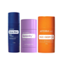 Three doTERRA body care stick containers on a plain white background: a dark blue Deep Blue stick on the left, a lavender Serenity stick in the center, and an orange Face + Body stick on the right.